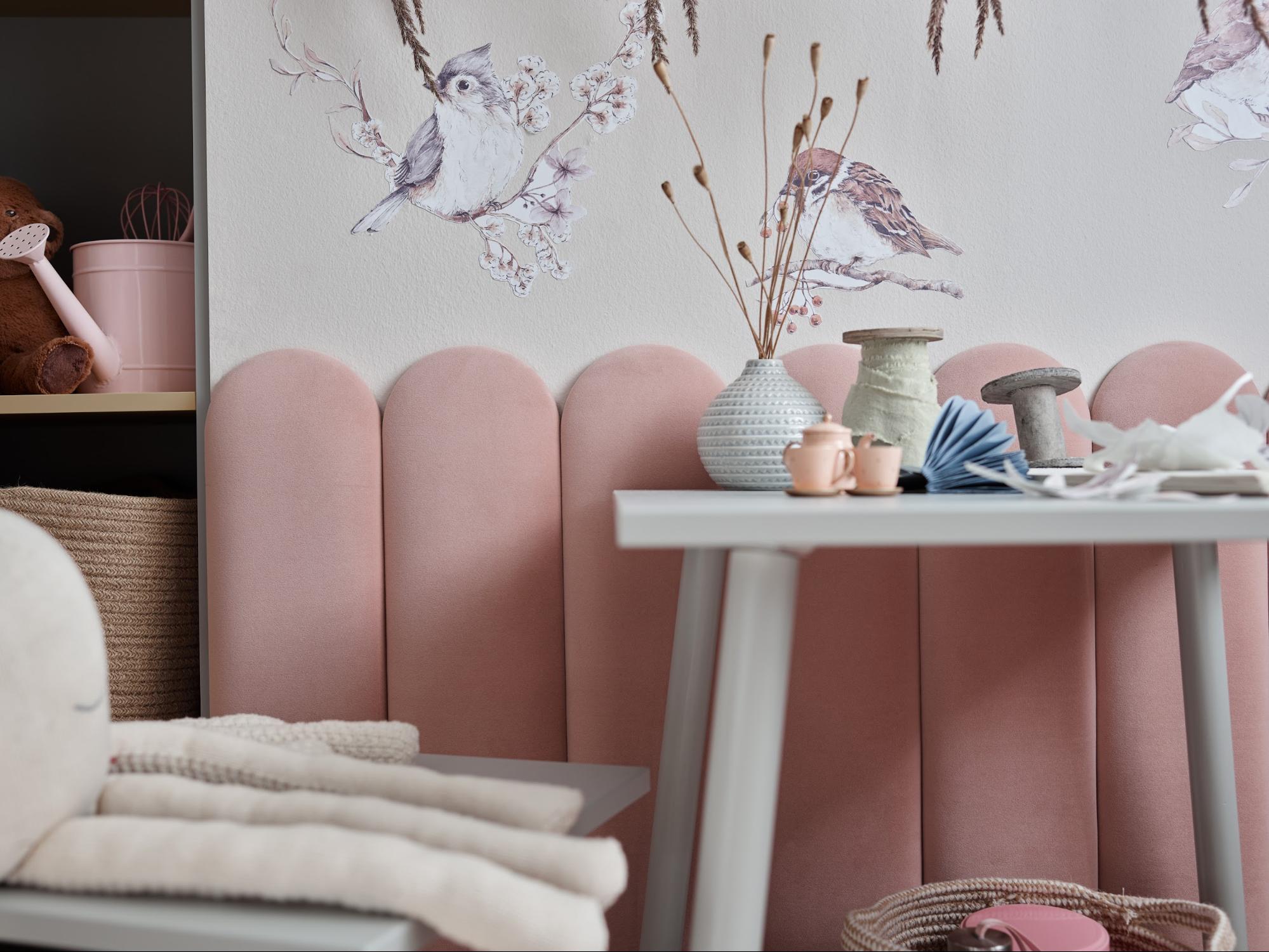 Przytulny kącik w pokoju dziecięcym z różową tapicerowaną ścianą i delikatnymi dekoracjami w postaci malunków ptaków.