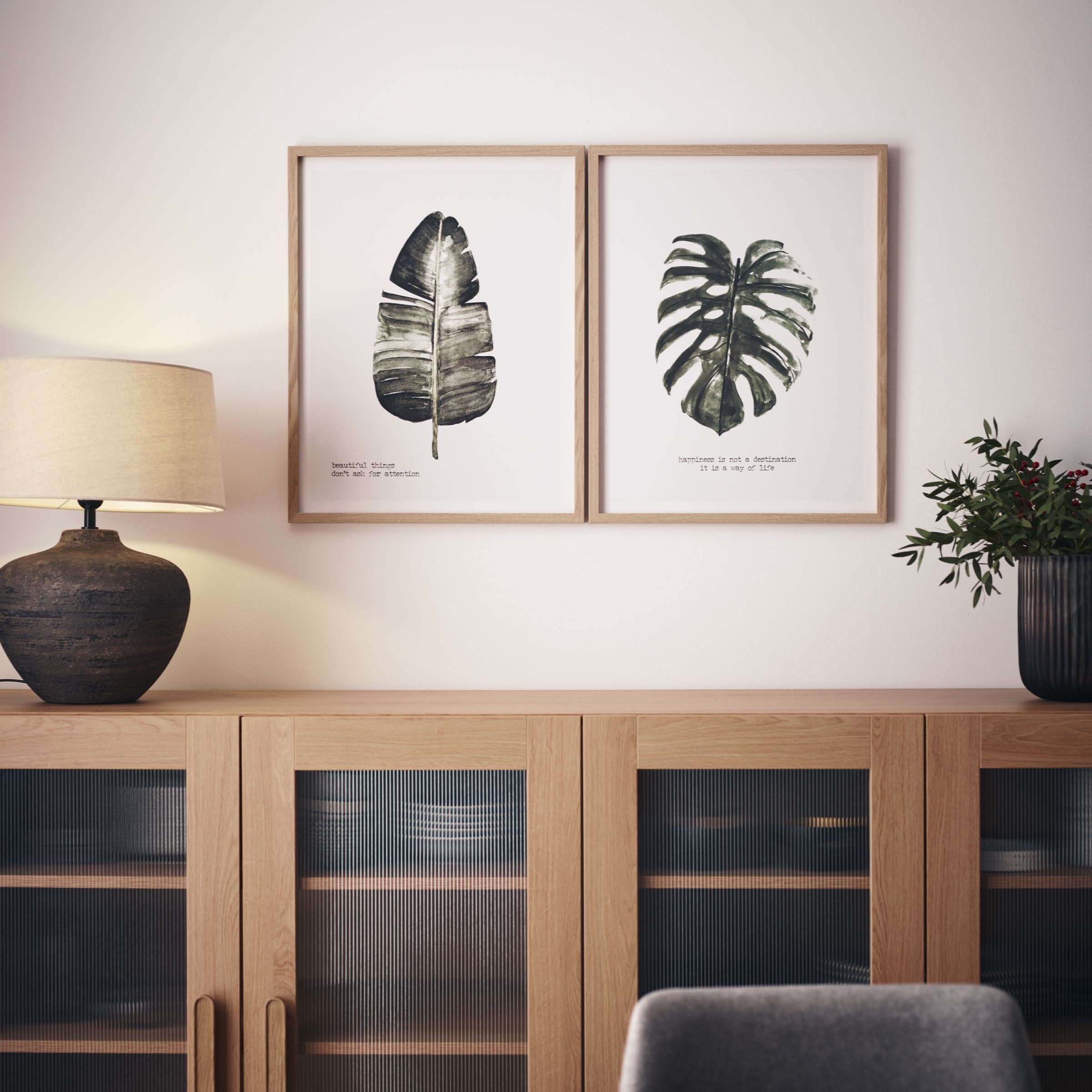 Grafiki z liśćmi nad komodą z lampą i rośliną