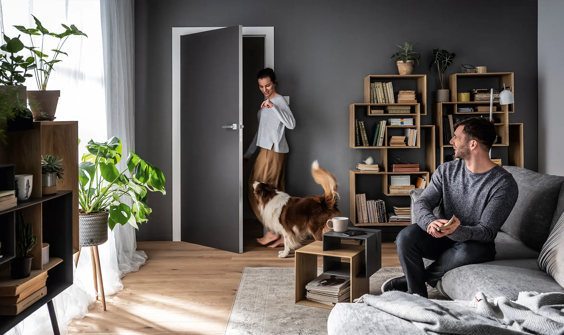 Ciemnoszare drzwi w salonie z grafitową ścianą, biblioteczką i dużą ilością roślin.