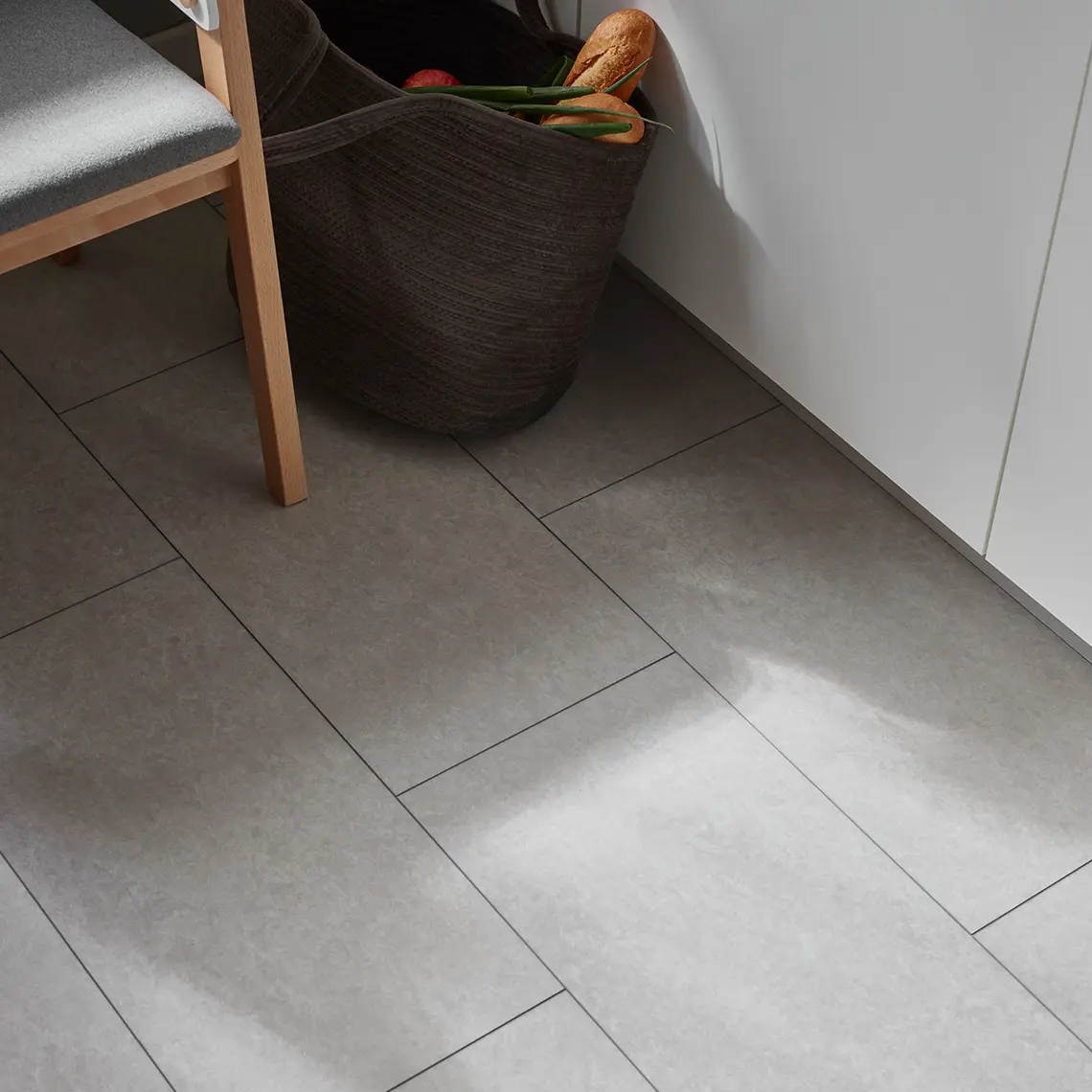 Zbliżenie na szarą podłogę imitującą beton w kuchni, obok drewnianego krzesła i kosza na zakupy.
