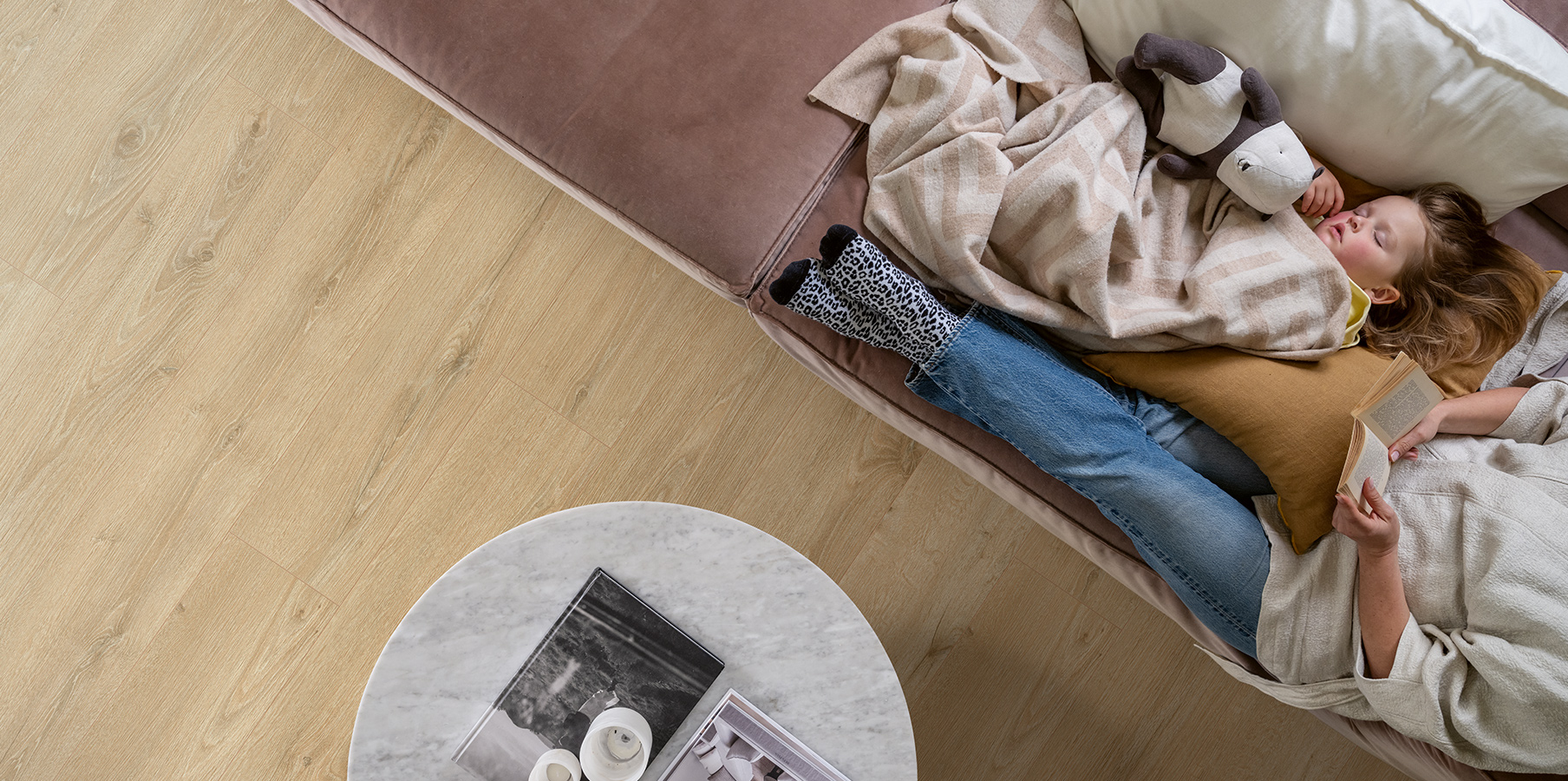 ciepła, drewniana podłoga i wygodna sofa, na której mama czyta książkę obok śpiącej córki.