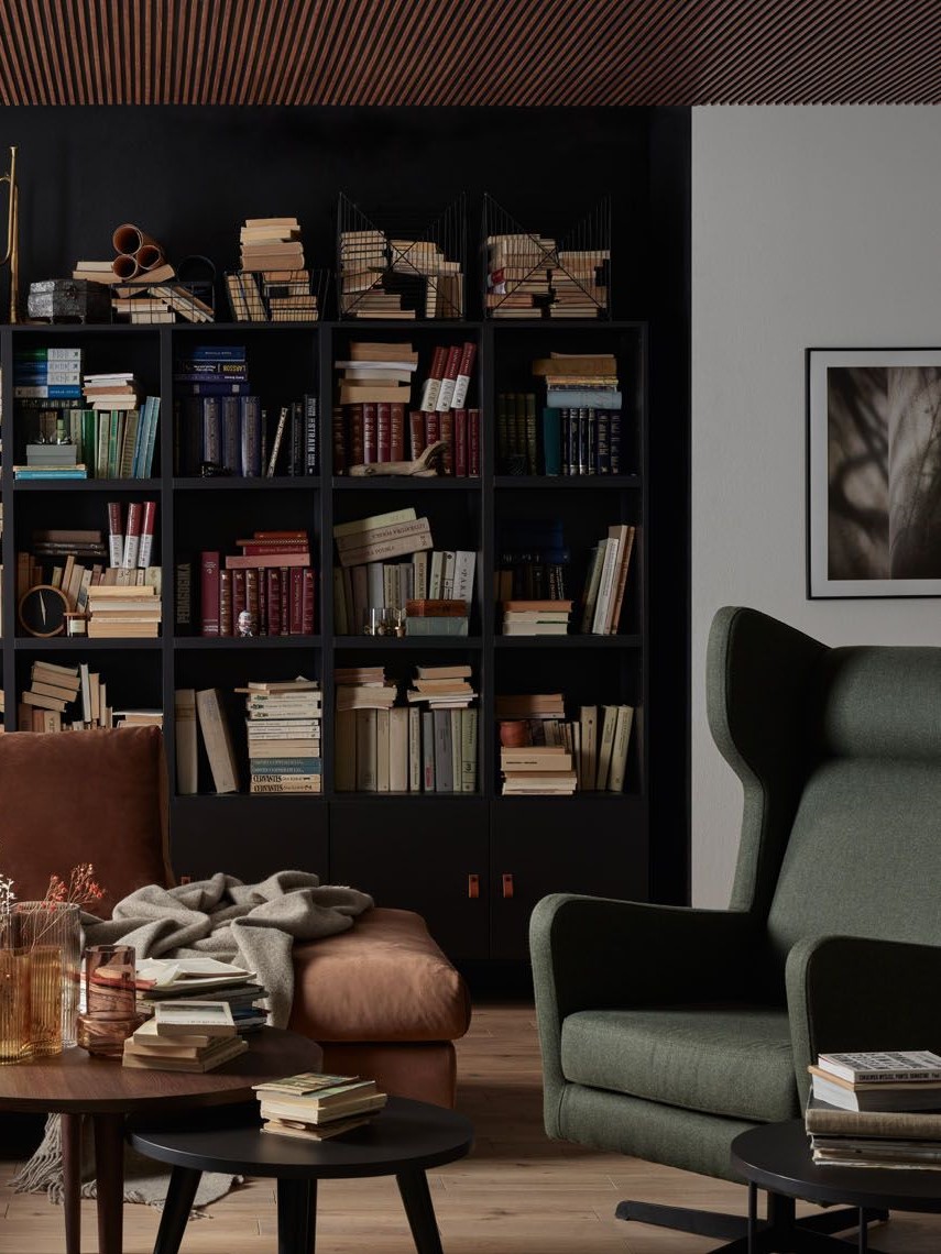 Elegancka biblioteka z czarnym regałem pełnym książek, ciemnozielonym fotelem i skórzaną sofą w ciepłym odcieniu brązu.