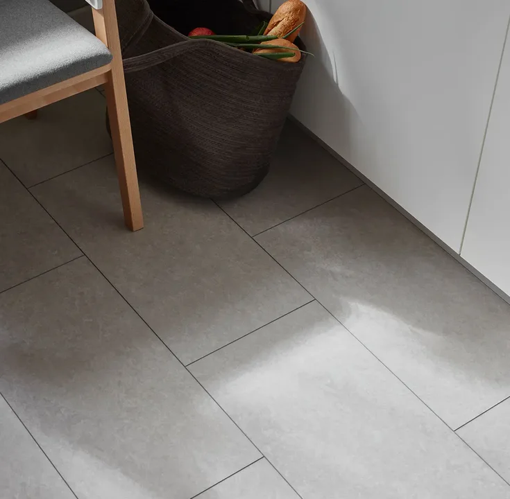 Zbliżenie na szarą podłogę imitującą beton w kuchni, obok drewnianego krzesła i kosza na zakupy.