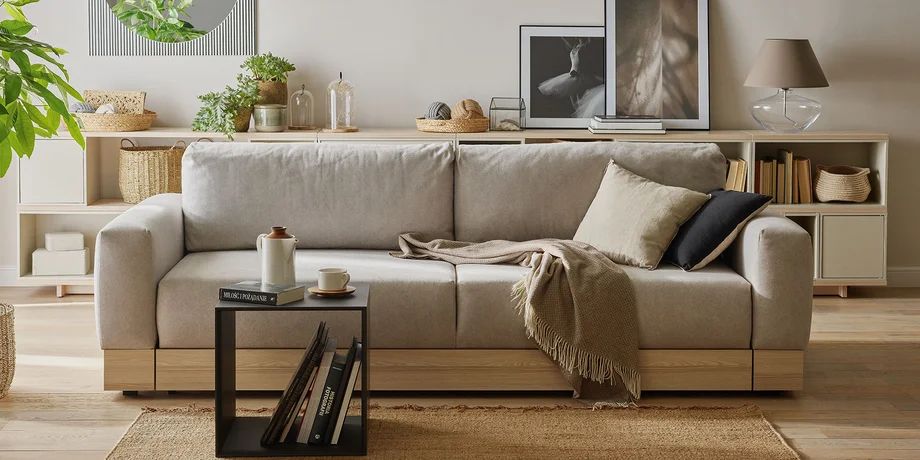 Przytulny salon z dużą, jasnoszarą sofą, drewnianymi i beżowymi dodatkami oraz roślinami, tworzący naturalną i harmonijną przestrzeń wypoczynkową.