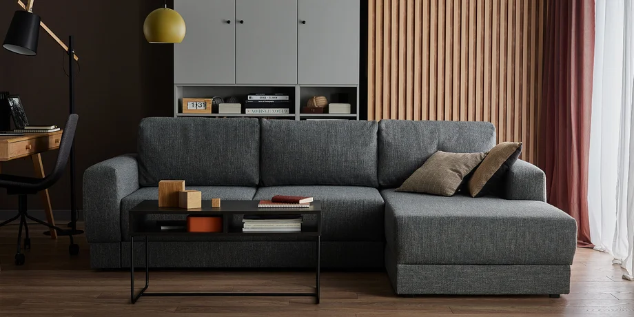 Szara, narożna sofa w eleganckim salonie z drewnianymi akcentami, stolikiem kawowym oraz dekoracjami w stylu nowoczesnym.