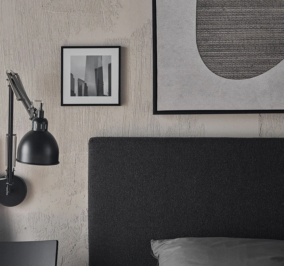 Elegancka sypialnia w monochromatycznych tonacjach z tapicerowanym zagłówkiem, industrialną lampką ścienną i minimalistycznymi grafikami na ścianie.
