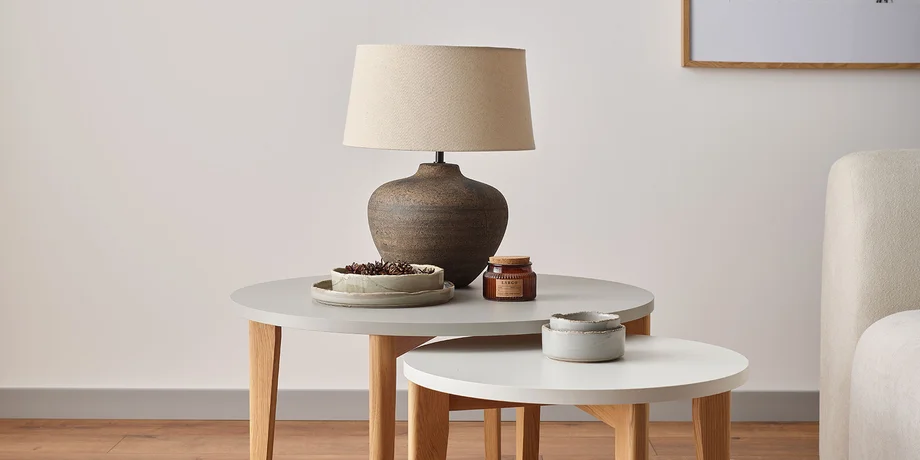 Stonowana lampa stołowa z ceramiczną podstawą i beżowym kloszem, ustawiona na zestawie okrągłych stolików,