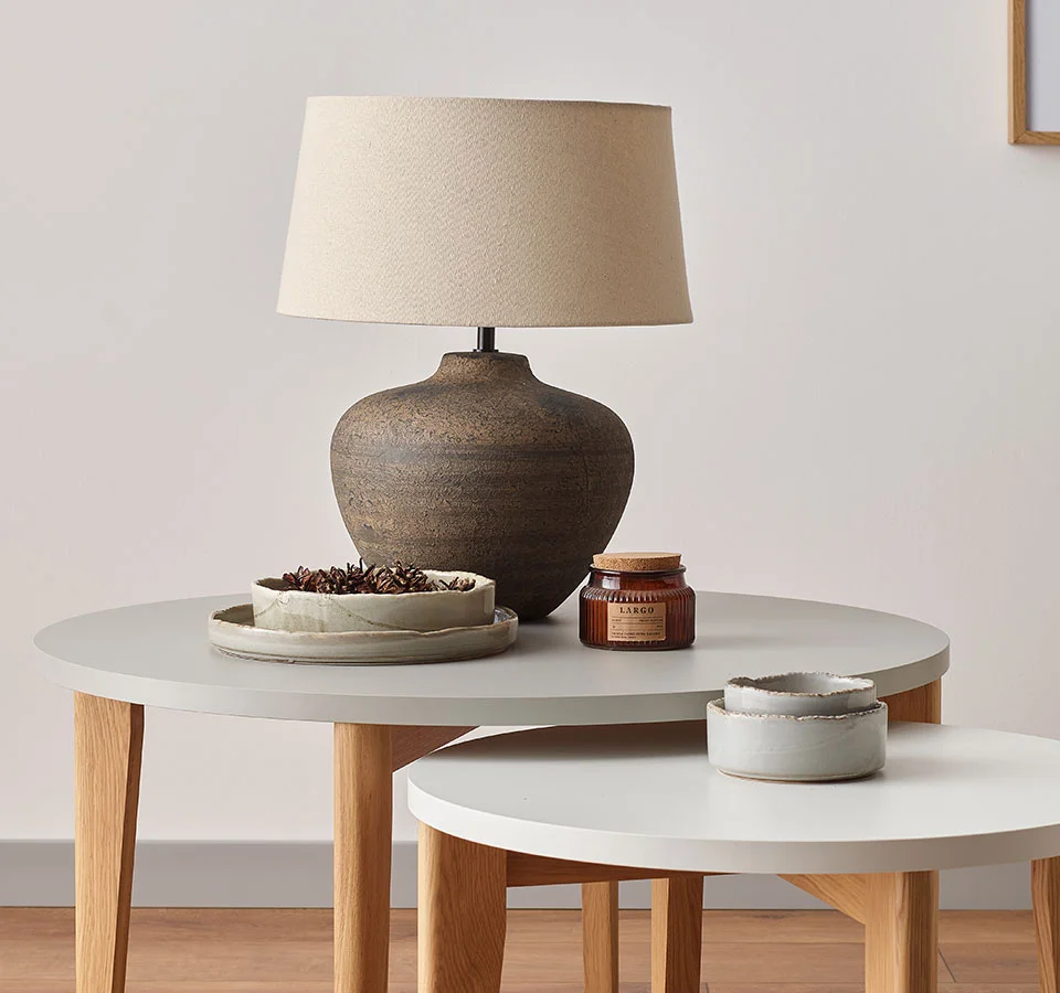 Stonowana lampa stołowa z ceramiczną podstawą i beżowym kloszem, ustawiona na zestawie okrągłych stolików,